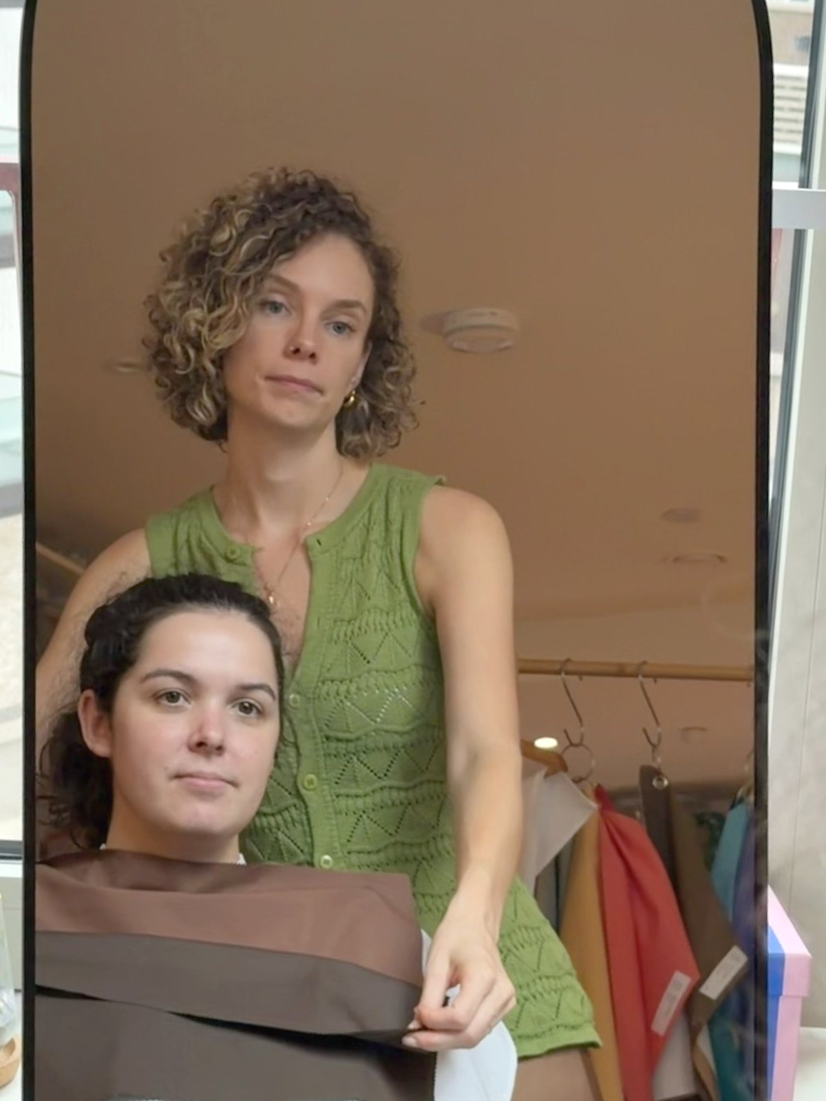 A client seated at the mirror with a drape held at her shoulders, while Shann compares it against the next one